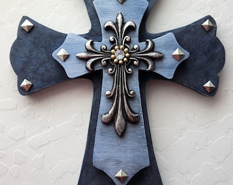 May include: A decorative cross with a layered design. The base is dark blue, with a light blue center. A silver-toned filigree cross with a central gem is overlaid. Silver studs are placed around the edges.