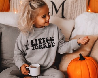 Op de afbeelding: Een jong kind draagt een grijs sweatshirt met de tekst "LITTLE TURKEY" in het zwart. Het kind zit op een bank met een pompoen voor zich en een kopje thee in de hand.