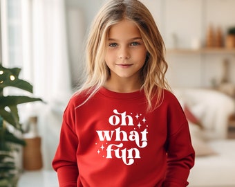 Può includere: Una felpa rossa con le parole "oh what fun" in bianco e stelle. La felpa è indossata da una persona giovane con i capelli biondi.