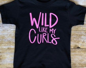 Op de afbeelding: Zwart T-shirt met roze opschrift "WILD LIKE MY CURLS".