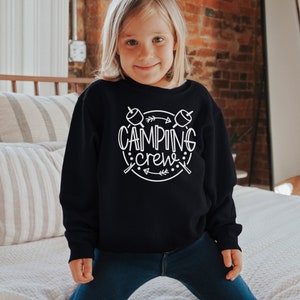 May include: A young person wearing a black sweatshirt with white text that says "Camping Crew" and a graphic of two marshmallows on sticks with arrows. The person is wearing blue jeans and is standing in front of a bed with white bedding.