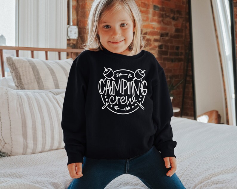 May include: A young person wearing a black sweatshirt with white text that says "Camping Crew" and a graphic of two marshmallows on sticks with arrows. The person is wearing blue jeans and is standing in front of a bed with white bedding.
