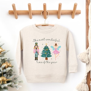 Puede incluir: Sudadera beige con la frase "The most wonderful time of the year". El diseño incluye un cascanueces, un árbol de Navidad y un hada. La sudadera está colgada de un perchero de madera.