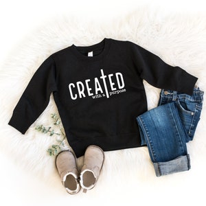 May include: Black sweatshirt with white text that reads "CREATED with a purpose" and a cross symbol. The sweatshirt is laid out on a white fluffy surface with a pair of brown boots and blue jeans.