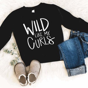 May include: Black long-sleeve shirt with white text that reads "WILD LIKE MY CURLS". The shirt is on a white fuzzy surface with a pair of brown boots and blue jeans.