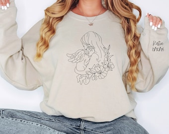 May include: A beige sweatshirt featuring a black line drawing of a mother holding a baby with angel wings, surrounded by flowers. The name "Katie" and the date "3/16/05" are printed on the right sleeve.