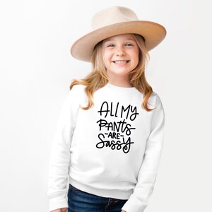 Może przedstawiać: Biała bluza z czarnym napisem "All My Pants -Are- Sassy". Młoda osoba nosi bluzę i brązowy kapelusz.