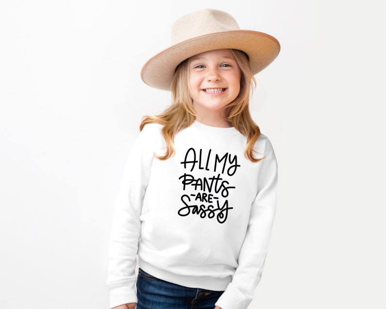 Może przedstawiać: Biała bluza z czarnym napisem "All My Pants -Are- Sassy". Młoda osoba nosi bluzę i brązowy kapelusz.