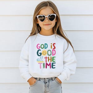 May include: A young person wearing heart-shaped sunglasses and a white sweatshirt. The sweatshirt has the text "GOD IS GOOD all THE TIME" in colorful letters. The person is also wearing blue jeans.