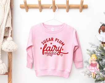 Può includere: Una felpa rosa chiaro con maniche lunghe, appesa a una gruccia bianca. La parte anteriore della felpa presenta il testo rosso "Sugar Plum Fairy magic" con stelle e cuori. La felpa è esposta su una parete bianca.