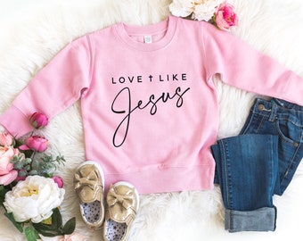 Peut inclure: Sweat-shirt rose pour enfant avec le texte "Love + Like Jesus" en écriture noire.