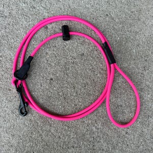 May include: A bright neon pink rope dog lead featuring a black metal swivel snap hook, a black sliding cord lock, and a black handle loop. The lead is constructed from a smooth, flexible cord material with black heat-shrink tubing accents.