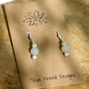 May include: A pair of dangle earrings with a light blue bead and a silver bead, suspended from a silver hook. The earrings are displayed on a brown card with a floral design and the text "True Friend Designs".