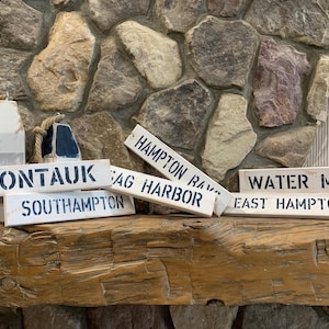 以下が含まれることがあります： ニューヨーク州ハンプトンにある、航海をテーマにした場所の名前が書かれた木製看板6枚セット。看板は白く塗られ、黒い文字で書かれています。看板に書かれている名前は、モントーク、サウサンプトン、サグハーバー、ハンプトンベイ、イーストハンプトン、ウォーターミルです。