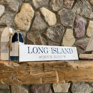 Può includere: Un cartello in legno bianco con il testo "LONG ISLAND" e le coordinate "40° 51' 5" N 73° 5' 57" W" in lettere nere. Il cartello è appoggiato su una trave di legno rustica con due decorazioni di boa nautica bianche e blu.