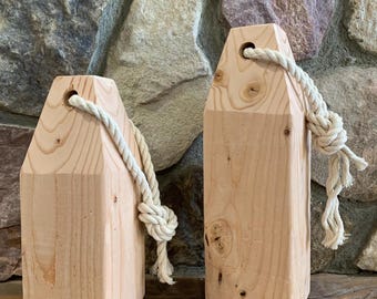 Peut inclure: Deux bouées en bois avec veinure naturelle. Chaque bouée a une forme rectangulaire avec une corde attachée à travers un trou en haut. Les cordes sont nouées. Les bouées sont exposées sur une surface en bois.