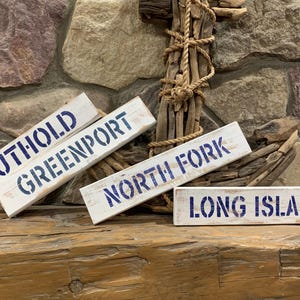 May include: Four white wooden signs with blue painted text. The signs read "Southold", "Greenport", "North Fork", and "Long Island".