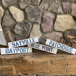 May include: Several white wooden signs with dark blue lettering are arranged on a wooden surface. The signs display the names of coastal towns: Sayville, Bayport, Blue Point, and Patchogue. The background features a stone wall.