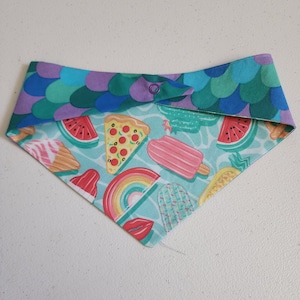 May include: A turquoise bandana with a summer-themed print featuring watermelon slices, popsicles, pizza slices, rainbows, and cacti. The bandana has a purple and green patterned border.