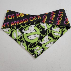 Puede incluir: Un bandana para perro de tela verde y negro con un patrón repetido de fantasmas blancos y limo negro. El bandana tiene un borde negro con el texto "I am not afraid of no ghosts" en un lado y "I am a host of ghosts" en el otro.