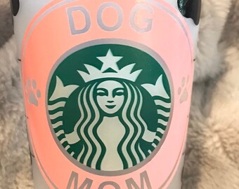 May include: Clear plastic tumbler with black paw prints and a pink and green logo that says "DOG MOM". The logo is a parody of a popular coffee shop logo. The tumbler is designed for dog lovers.