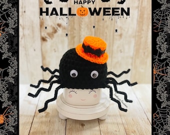 May include: A black crocheted spider topper with googly eyes and an orange top hat, resting on a white ceramic mug with a printed face. The background features the text HAPPY HALLOWEEN with a pumpkin icon and black bats, spiders, and webs.