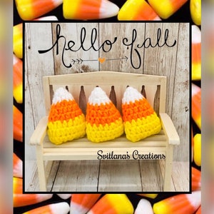 May include: A miniature light-colored wooden bench featuring three crocheted candy corn decorations in white, orange, and yellow. The text "hello fall" and "Svitlana's Creations" is printed on the image. The background displays candy corn.