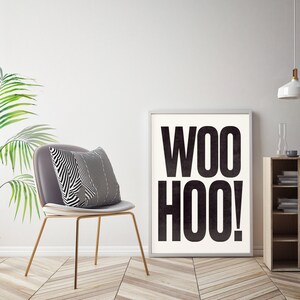 May include: A black and white poster with the text "WOO HOO!" in bold, large font. The poster is framed in a white frame and is hanging on a white wall.