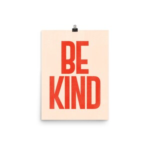 Puede incluir: Un p&oacute;ster beige con el texto "BE KIND" en letras rojas. Las letras son negritas y de bloque.