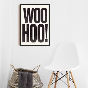 May include: A black and white framed print with the text "WOO HOO!" hanging on a white wall. A white chair with wooden legs and a woven basket with a gray blanket are in the foreground.
