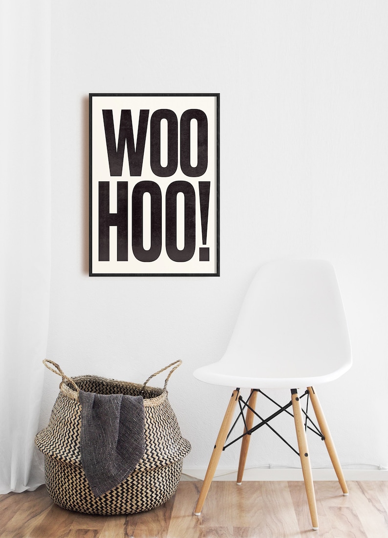 May include: A black and white framed print with the text "WOO HOO!" hanging on a white wall. A white chair with wooden legs and a woven basket with a gray blanket are in the foreground.
