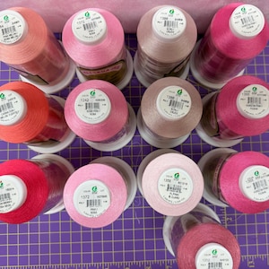Könnte beinhalten: Eine Sammlung von Garnrollen in verschiedenen Rosatönen, von hellem Rosa bis zu tiefem Magenta. Die Spulen sind auf einem lila Raster angeordnet und zeigen eine Reihe von Farben für Näh- und Bastelprojekte.
