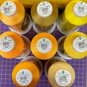 Puede incluir: Surtido de diez carretes de hilo en varios tonos de amarillo y naranja. Cada carrete está etiquetado con la marca "Iris" e incluye el número de color, el número de lote y el nombre del color en inglés y español. Los carretes están dispuestos en círculo sobre una superficie a cuadros blancos y morados.