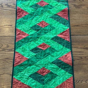 May include: A green and red quilted table runner with a geometric pattern. The runner has a dark green border.