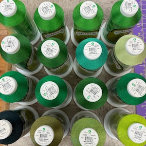 Op de afbeelding: Een verzameling van 12 spoelen groene draad, elk met een wit label en een kleurnummer. De labels bevatten de kleurnaam en het draadmateriaal, dat polyester is.