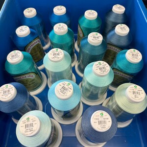 May include: A close-up of a collection of spools of thread in various shades of blue and green. The spools are arranged in a blue plastic container. The spools are labeled with the brand name "POLYSEDA IRIS" and the color number.