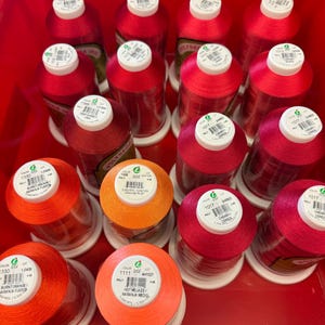 May include: A collection of spools of red, orange, and pink embroidery thread. The spools are labeled with color numbers and names, such as "Burnt Orange" and "Cherry".