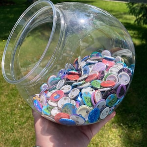 May include: A clear glass jar filled with colourful round buttons. The buttons have various designs, including geometric shapes, pixelated images, and text. The jar is sitting on a person's hand.