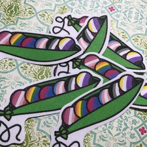 May include: Glittery stickers featuring green pea pods with colorful, striped peas inside. The peas display various LGBTQ+ pride flag designs, including rainbow, transgender, and non-binary flags. The stickers have a white border.