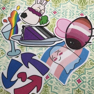 Puede incluir: Una colección de pegatinas coloridas con varios diseños. Incluye un conejo de dibujos animados con un collar morado, una rebanada de pastel morado, una bebida arcoíris, una mariquita con alas rosas y marrones, un fantasma con una bandera transgénero y flechas con un corazón.