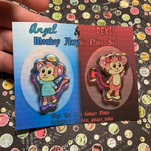 May include: A set of two iridescent acrylic pins featuring a monkey dressed as an angel and a monkey dressed as a devil. The pins are on a card with the text "Angel & Devil Monkey Acrylic Pins Set".