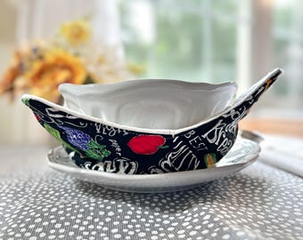 May include: A white ceramic bowl with a black and white fabric bowl cozy. The cozy has a chalkboard-style design with the words "Best Mill" and "Pepper" and images of vegetables and fruit.
