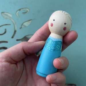 Puede incluir: Una mu&ntilde;eca de madera pintada a mano con un vestido azul y detalles de purpurina. La mu&ntilde;eca tiene una cara marr&oacute;n claro con mejillas rosadas y ojos negros.