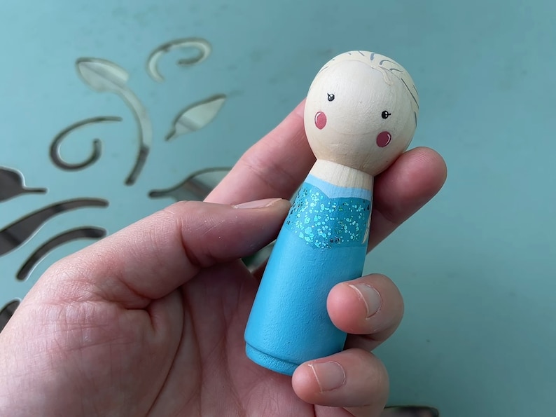 Puede incluir: Una mu&ntilde;eca de madera pintada a mano con un vestido azul y detalles de purpurina. La mu&ntilde;eca tiene una cara marr&oacute;n claro con mejillas rosadas y ojos negros.
