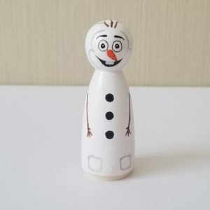 Puede incluir: Una figurita de mu&ntilde;eco de nieve de madera blanca con brazos de rama marr&oacute;n, botones negros y una nariz de zanahoria. El mu&ntilde;eco de nieve tiene una cara pintada con ojos negros, una boca negra y una sonrisa negra.
