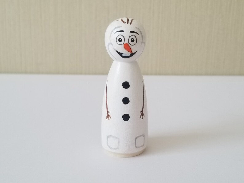 Puede incluir: Una figurita de mu&ntilde;eco de nieve de madera blanca con brazos de rama marr&oacute;n, botones negros y una nariz de zanahoria. El mu&ntilde;eco de nieve tiene una cara pintada con ojos negros, una boca negra y una sonrisa negra.