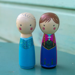 Puede incluir: Dos mu&ntilde;ecas de madera con clavijas vestidas como los personajes Elsa y Anna de la pel&iacute;cula Frozen. Elsa lleva un vestido azul con purpurina y Anna lleva un vestido rojo y azul con una trenza.
