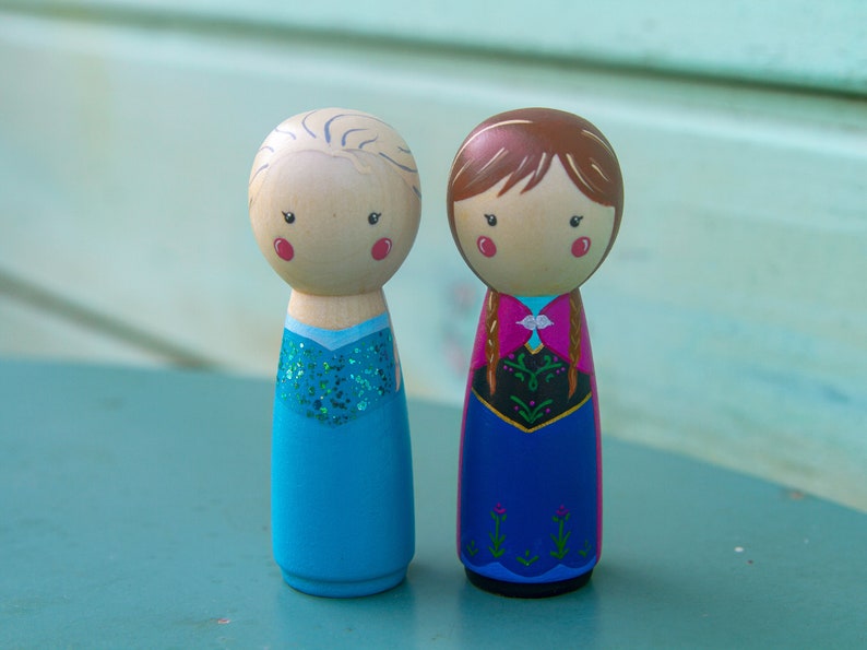 Puede incluir: Dos mu&ntilde;ecas de madera con clavijas vestidas como los personajes Elsa y Anna de la pel&iacute;cula Frozen. Elsa lleva un vestido azul con purpurina y Anna lleva un vestido rojo y azul con una trenza.
