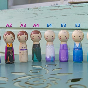 Puede incluir: Ocho mu&ntilde;ecas de madera con clavijas pintadas para parecerse a personajes de la pel&iacute;cula Frozen. Las mu&ntilde;ecas est&aacute;n dispuestas en una fila de izquierda a derecha, etiquetadas de A1 a A4 y de E1 a E4. Las mu&ntilde;ecas llevan diferentes atuendos, incluyendo un vestido azul con una capa rosa, un atuendo negro y marr&oacute;n, un vestido beige con detalles dorados, un vestido verde con detalles turquesa, un vestido blanco con detalles azules, un vestido morado con detalles azules, un vestido azul con detalles blancos y un vestido azul con detalles de purpurina.