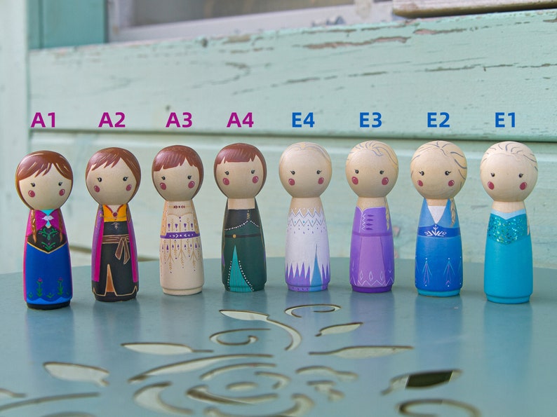 Puede incluir: Ocho mu&ntilde;ecas de madera con clavijas pintadas para parecerse a personajes de la pel&iacute;cula Frozen. Las mu&ntilde;ecas est&aacute;n dispuestas en una fila de izquierda a derecha, etiquetadas de A1 a A4 y de E1 a E4. Las mu&ntilde;ecas llevan diferentes atuendos, incluyendo un vestido azul con una capa rosa, un atuendo negro y marr&oacute;n, un vestido beige con detalles dorados, un vestido verde con detalles turquesa, un vestido blanco con detalles azules, un vestido morado con detalles azules, un vestido azul con detalles blancos y un vestido azul con detalles de purpurina.
